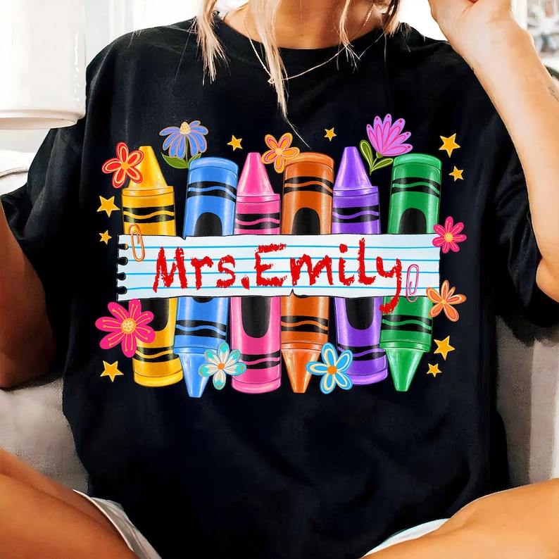May include: Black t-shirt featuring a colorful crayon design with the text "Mrs. Emily". The design includes yellow, blue, pink, orange, purple, and green crayons, along with flowers and stars.