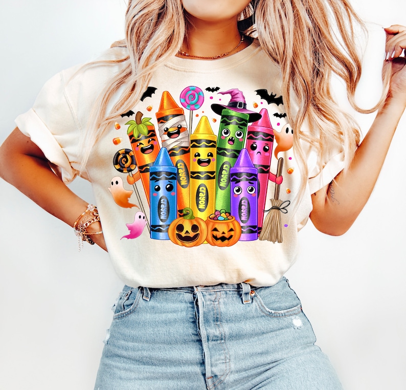 May include: Cream-colored t-shirt featuring a colorful Halloween-themed crayon design. The design includes smiling crayons dressed as Halloween characters, pumpkins, ghosts, and bats. The word "CRAYOLA" is visible on the crayons.