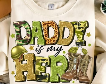 Könnte beinhalten: Ein weißer Pullover mit dem Text "Daddy is my Hero" in Grün, Gold und Tarnmuster. Der Text ist mit Glitzer und Sternen verziert. Es gibt zwei braune Stiefel mit grünen Akzenten und einen grünen Helm mit Tarnmuster.
