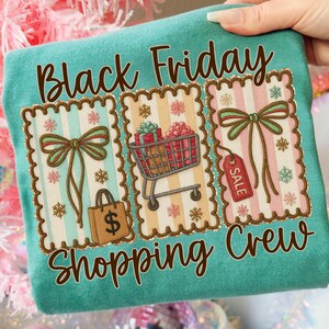 May include: Teal t-shirt with the words "Black Friday Shopping Crew" in brown script. The shirt features three decorative panels with bows, a shopping cart, a shopping bag, and a sale tag.