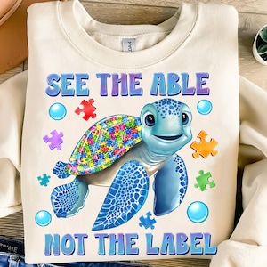 May include: A cream-colored sweatshirt with a blue sea turtle with a puzzle piece shell. The turtle is surrounded by puzzle pieces and bubbles. The text "See the Able Not the Label" is printed on the sweatshirt.