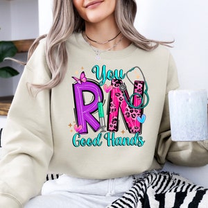 May include: A beige sweatshirt with a graphic design that says "You RN Good Hands". The graphic features a purple "R", a pink "N" with a leopard print, a stethoscope, a butterfly, and a syringe.