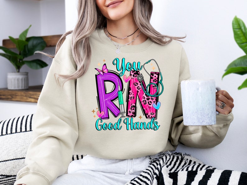 May include: A beige sweatshirt with a graphic design that says "You RN Good Hands". The graphic features a purple "R", a pink "N" with a leopard print, a stethoscope, a butterfly, and a syringe.
