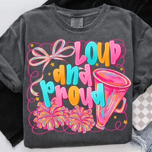 May include: Dark gray t-shirt with the words "LOUD and PROUD" in bright pink, blue, and orange. The design includes a megaphone, pom-poms, and a bow. The shirt is a crew neck with long sleeves.
