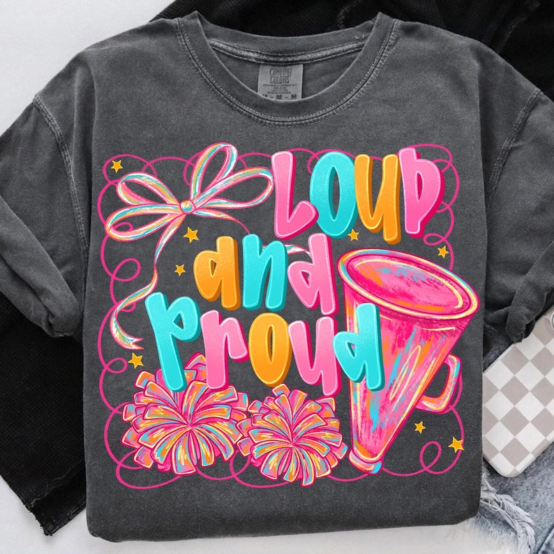 May include: Dark gray t-shirt with the words "LOUD and PROUD" in bright pink, blue, and orange. The design includes a megaphone, pom-poms, and a bow. The shirt is a crew neck with long sleeves.