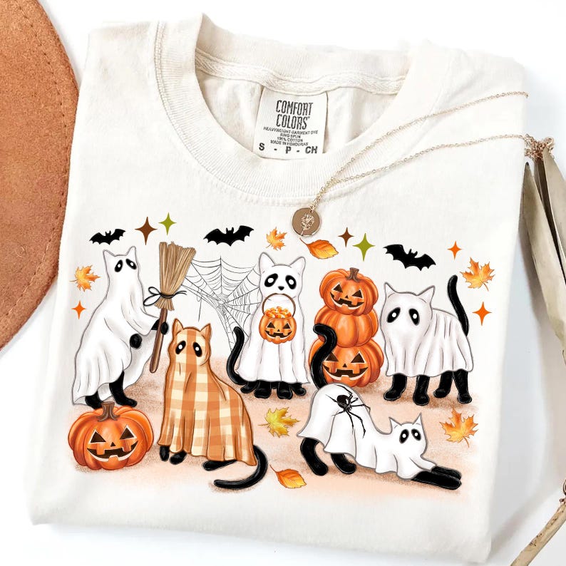 May include: A cream-coloured t-shirt with a Halloween graphic. The design features cartoon cats dressed as ghosts, pumpkins, and a cat in a plaid costume. The shirt also has bats, a spider web, and autumn leaves. The shirt has the Comfort Colors brand label.