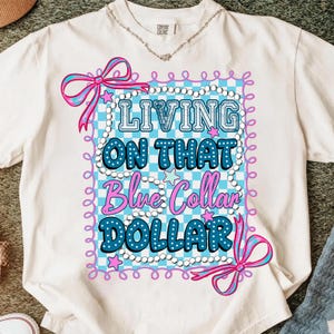 Pu&ograve; includere: T-shirt bianco con la scritta "LIVING ON THAT Blue Collar DOLLAR" in blu e bianco. Il disegno &egrave; incorniciato da un motivo a scacchi azzurro e bianco, fiocchi rosa e stelle. Visibile anche una collana d'argento.