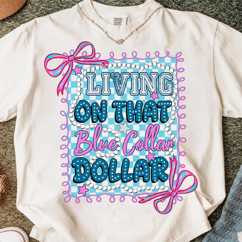 Pu&ograve; includere: T-shirt bianco con la scritta "LIVING ON THAT Blue Collar DOLLAR" in blu e bianco. Il disegno &egrave; incorniciato da un motivo a scacchi azzurro e bianco, fiocchi rosa e stelle. Visibile anche una collana d'argento.