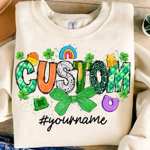 May include: A white sweatshirt with a green, orange, and black "CUSTOM" design with a green bow and shamrock accents. The text "#yourname" is below the design.