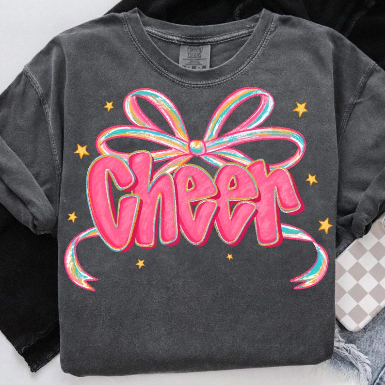 May include: Dark gray long-sleeve shirt with a pink "Cheer" graphic. The word "Cheer" is in a bubble letter style, with a colorful bow and yellow stars. The shirt is made of a soft, comfortable material.