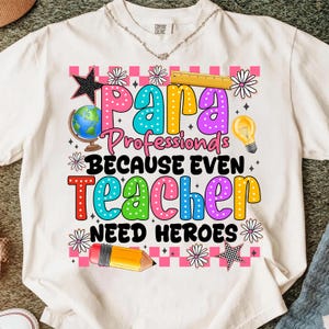 May include: Cream-colored t-shirt with a colorful graphic design. The design includes the words "Para Professionals Because Even Teacher Need Heroes" with school-themed illustrations like a globe, pencil, and lightbulb.