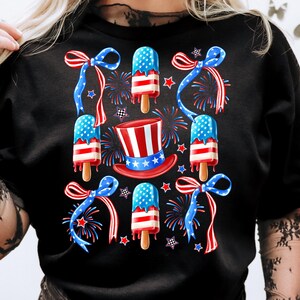 Puede incluir: Camiseta negra con un tema del 4 de julio en rojo, blanco y azul. El dise&ntilde;o presenta paletas de hielo, lazos y fuegos artificiales.