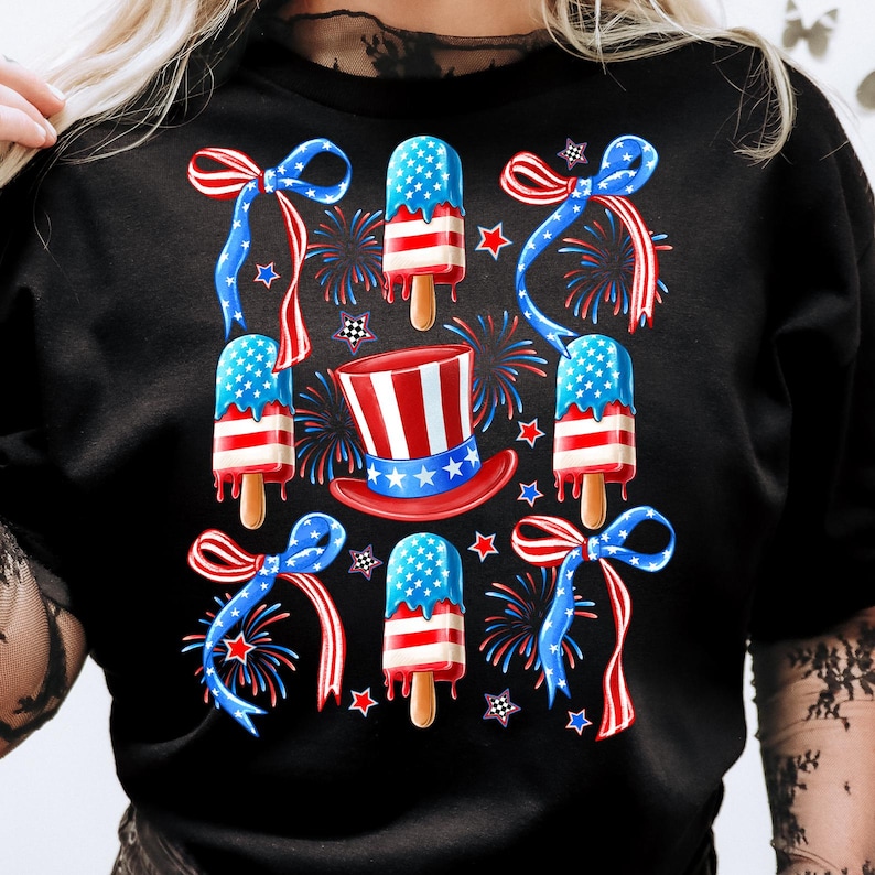 Puede incluir: Camiseta negra con un tema del 4 de julio en rojo, blanco y azul. El dise&ntilde;o presenta paletas de hielo, lazos y fuegos artificiales.