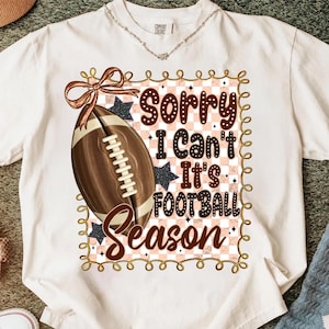 May include: Cream-colored t-shirt with a football graphic and the text "Sorry I Can't It's Football Season." The design includes a brown football, a bow, and stars. The shirt has a round neckline and short sleeves.