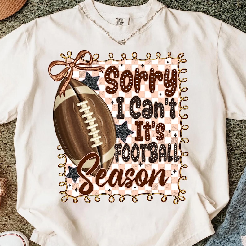 May include: Cream-colored t-shirt with a football graphic and the text "Sorry I Can't It's Football Season." The design includes a brown football, a bow, and stars. The shirt has a round neckline and short sleeves.