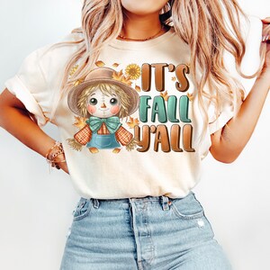 Op de afbeelding: Een wit T-shirt met een cartoon-vogelverschrikker en de tekst "It's Fall Y'all".