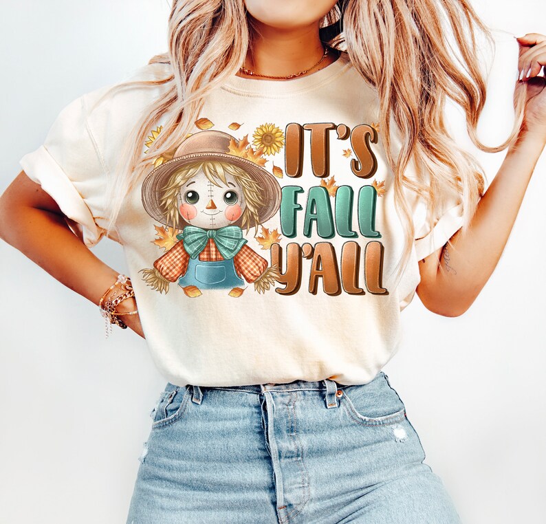 Op de afbeelding: Een wit T-shirt met een cartoon-vogelverschrikker en de tekst "It's Fall Y'all".