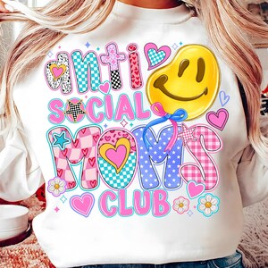 May include: White sweatshirt with a pink, blue, and yellow graphic design that says "Anti Social Moms Club". The graphic includes a smiley face, stars, hearts, and checkered patterns.