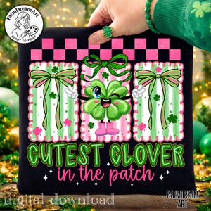 May include: Black t-shirt featuring a cute cartoon clover with a winking eye, pink boots, and white gloves. The clover is set against a pink and green striped background with a green bow. The text "Cutest Clover in the Patch" is displayed in green.