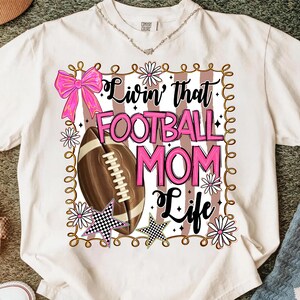 May include: Cream-colored t-shirt with a graphic that reads "Livin' that FOOTBALL MOM Life" in pink and black. The design includes a football, a pink bow, daisies, and star accents. The shirt is a Comfort Colors brand.