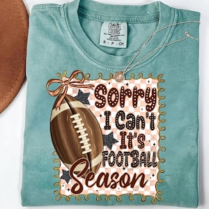 May include: A teal Comfort Colors t-shirt with a football graphic and the text "Sorry I Can't It's Football Season." The graphic includes a brown football, a brown bow, and black stars. A silver necklace is draped over the shirt.