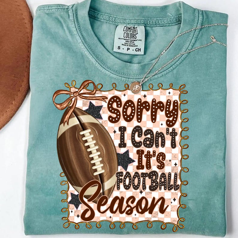 May include: A teal Comfort Colors t-shirt with a football graphic and the text "Sorry I Can't It's Football Season." The graphic includes a brown football, a brown bow, and black stars. A silver necklace is draped over the shirt.