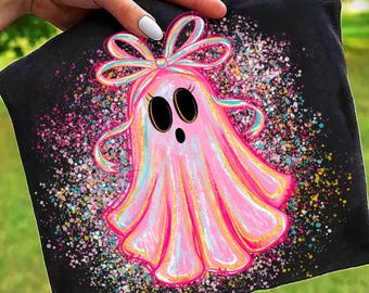 May include: Black t-shirt with a pink ghost design. The ghost has a bow, large black eyes, and a surprised expression. The design is embellished with colourful speckles and gold accents, creating a whimsical and festive appearance.
