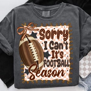 May include: Dark gray sweatshirt with a football graphic. The graphic includes a brown football, a bow, and the text "Sorry I can't It's Football Season" in brown and white. The design has a checkered background and star accents.