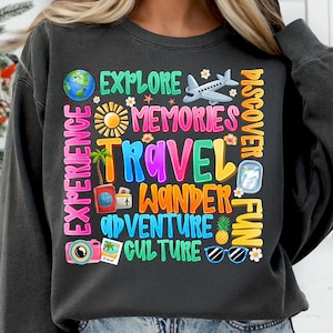 May include: A dark gray sweatshirt with a colorful graphic design featuring words related to travel, such as "explore", "memories", "travel", "wander", "adventure", "culture", and "fun". The graphic also includes various travel-related icons, including a plane, a suitcase, a camera, sunglasses, and a pineapple.