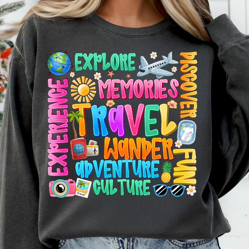 May include: A dark gray sweatshirt with a colorful graphic design featuring words related to travel, such as "explore", "memories", "travel", "wander", "adventure", "culture", and "fun". The graphic also includes various travel-related icons, including a plane, a suitcase, a camera, sunglasses, and a pineapple.