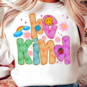 May include: White sweatshirt with the words "be kind" in colorful, whimsical lettering. The design includes a rainbow, smiley faces, flowers, and a ladybug. The sweatshirt has a crew neck and long sleeves.