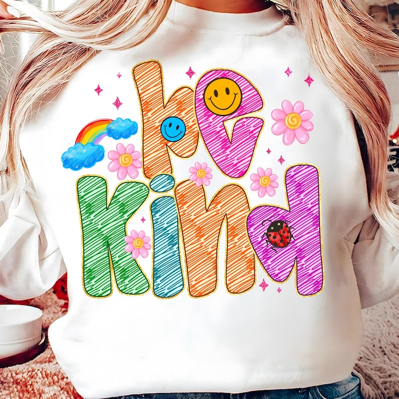 May include: White sweatshirt with the words "be kind" in colorful, whimsical lettering. The design includes a rainbow, smiley faces, flowers, and a ladybug. The sweatshirt has a crew neck and long sleeves.