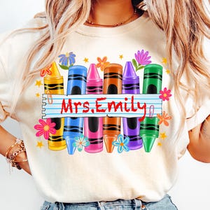 May include: Cream-colored sweatshirt featuring a colorful crayon design with the text "Mrs. Emily". The design includes crayons in yellow, blue, pink, brown, purple, and green, along with flowers and stars.
