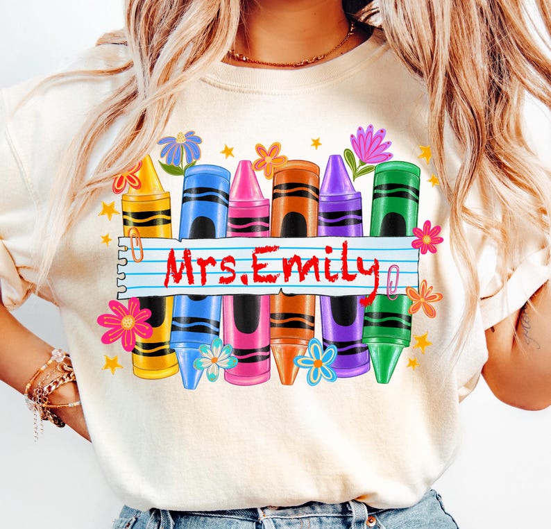 May include: Cream-colored sweatshirt featuring a colorful crayon design with the text "Mrs. Emily". The design includes crayons in yellow, blue, pink, brown, purple, and green, along with flowers and stars.