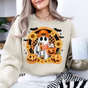 Peut inclure: Sweat-shirt cr&egrave;me avec un motif sur le th&egrave;me d'Halloween. Le motif comprend un fant&ocirc;me de dessin anim&eacute; portant un bonnet orange, tenant une boisson et un seau de citrouille, entour&eacute; de tournesols, de chauves-souris et de citrouilles. Le motif est dans un cadre circulaire.