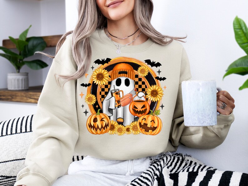 Peut inclure: Sweat-shirt cr&egrave;me avec un motif sur le th&egrave;me d'Halloween. Le motif comprend un fant&ocirc;me de dessin anim&eacute; portant un bonnet orange, tenant une boisson et un seau de citrouille, entour&eacute; de tournesols, de chauves-souris et de citrouilles. Le motif est dans un cadre circulaire.