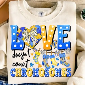 May include: A cream-colored sweatshirt with a blue and yellow graphic design. The graphic features the word "LOVE" in large, stylized letters with polka dots. The letters are decorated with puzzle pieces and bows. Below the word "LOVE" is the text "doesn't count CHROMOSOMES".