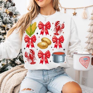 K&ouml;nnte beinhalten: Wei&szlig;er Pullover mit einem festlichen Design mit roten Schleifen, Maiskolben, Kartoffeln, Krebsen und einem Topf mit roter Sauce.