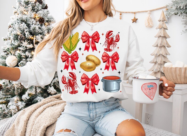 K&ouml;nnte beinhalten: Wei&szlig;er Pullover mit einem festlichen Design mit roten Schleifen, Maiskolben, Kartoffeln, Krebsen und einem Topf mit roter Sauce.