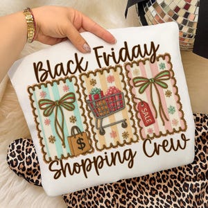 May include: White sweatshirt with the text "Black Friday Shopping Crew." The design features three rectangular images with bows, shopping carts, and gift boxes. The sweatshirt is decorated with snowflakes and a shopping bag.