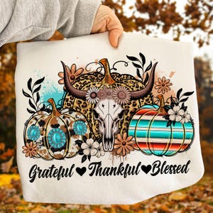 Puede incluir: Gr&aacute;fico color crema con tres calabazas con dise&ntilde;os florales y del suroeste. La calabaza central tiene una calavera con cuernos. El texto "Grateful, Thankful, Blessed" est&aacute; debajo.