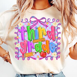 Può includere: Maglietta a maniche lunghe color crema con un design grafico colorato per la terza elementare. Il design include le parole "third grade" nei colori dell'arcobaleno, un righello, una matita, una mela e elementi floreali.