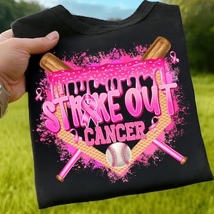 May include: Black t-shirt with a pink graphic that says "Strike Out Cancer." The design includes a baseball, crossed baseball bats, and pink ribbons. The shirt is held up against a green background.