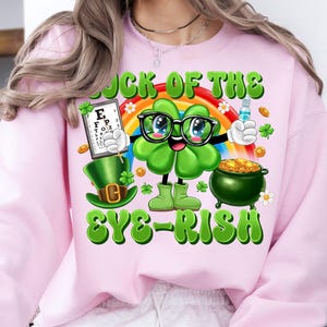 May include: Pink sweatshirt with a green shamrock cartoon character wearing glasses and holding an eye chart. The text "Luck of the Eye-rish" is printed on the shirt.