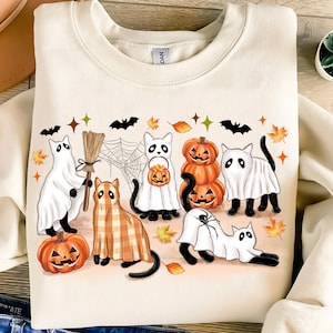 May include: Cream-coloured sweatshirt with a Halloween graphic. The design features cartoon cats dressed as ghosts, pumpkins, and a cat in a plaid costume. The design also includes bats, spiderwebs, and autumn leaves.