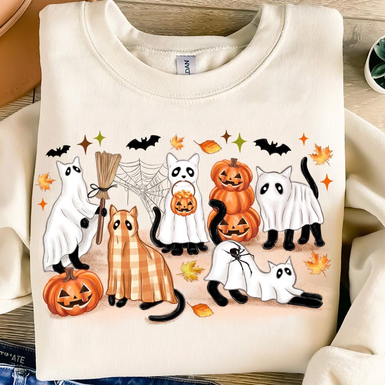 May include: Cream-coloured sweatshirt with a Halloween graphic. The design features cartoon cats dressed as ghosts, pumpkins, and a cat in a plaid costume. The design also includes bats, spiderwebs, and autumn leaves.