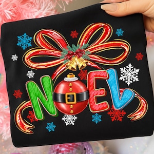 Pu&ograve; includere: Felpa nera con un design festivo. Presenta la parola "NOEL" in lettere verdi, rosse e blu, un ornamento rosso con cintura nera e un fiocco rosso con bordi dorati. Include fiocchi di neve e campane.