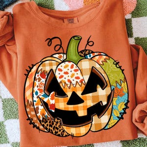 May include: An orange Comfort Colors sweatshirt with a patchwork pumpkin design. The pumpkin has a carved face and is made up of different patterns, including plaid, floral, and leopard print. The sweatshirt is a fall-themed item.