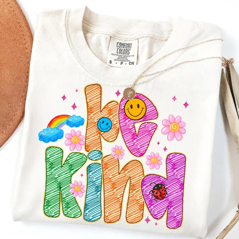 May include: Cream-colored t-shirt with the words "be kind" in colorful, scribbled letters. The design includes a rainbow, flowers, smiley faces, and a ladybug. A gold necklace with a pendant is draped over the shirt.