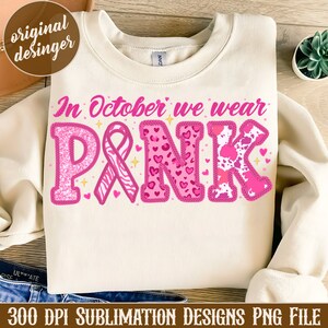 May include: A white sweatshirt with the text "In October we wear PINK" in pink letters. The letters are decorated with pink glitter, hearts, and animal print patterns. A pink ribbon is included in the design.